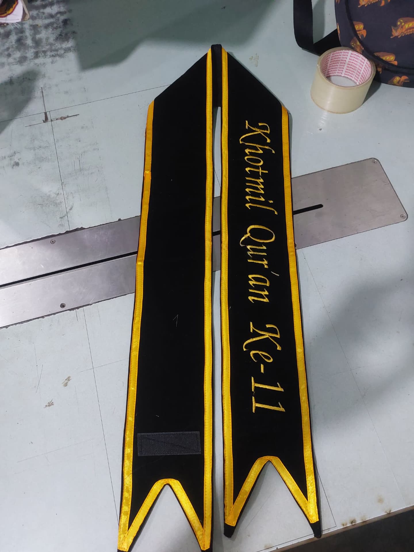 Selempang gelap dengan bordir tulisan dan garis kuning.